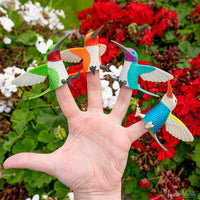 four colorful hummingbird finger puppets on a hand agains flowers