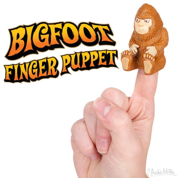 Bigfoot finger puppet sitting on raised index finger, with colorful text "BIGFOOT FINGER PUPPET" above. Brown, furry creature with large feet and expressive face. Fun, playful toy for Sasquatch enthusiasts.