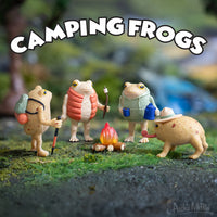 Four funny Camping Frogs figurines gathered around a campfire enjoying their outdoor adventure.