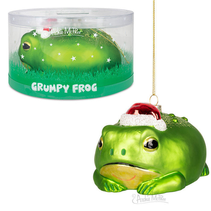 Hand-blown glass Christmas ornament featuring a grumpy green frog wearing a Santa hat, displayed in a clear cylindrical container with grass-like base and starry background, perfect for adding whimsical holiday cheer to your tree or table decor