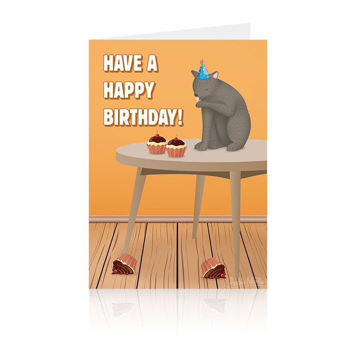 Grey cat wearing birthday hat sitting on table with cupcakes, cleaning itself. Text reads "Have A Happy Birthday!" Wooden floor with fallen cake pieces. Humorous greeting card design.