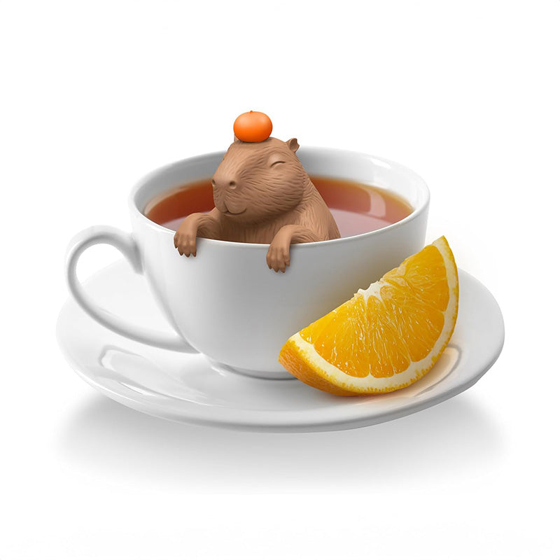 A brown capybara-shaped tea infuser with a small orange on its head sitting in a white cup of tea on a saucer with a slice of orange