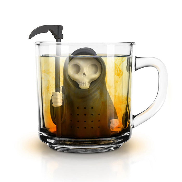 Grim Steeper tea infuser shaped like Death, with skull face and scythe, steeping in clear glass mug filled with golden tea. Unique and humorous kitchen accessory for tea lovers and Halloween enthusiasts.