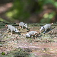 Four small grey plastic figures called Itty Bitty Possums are resting on a weathered wooden surface.