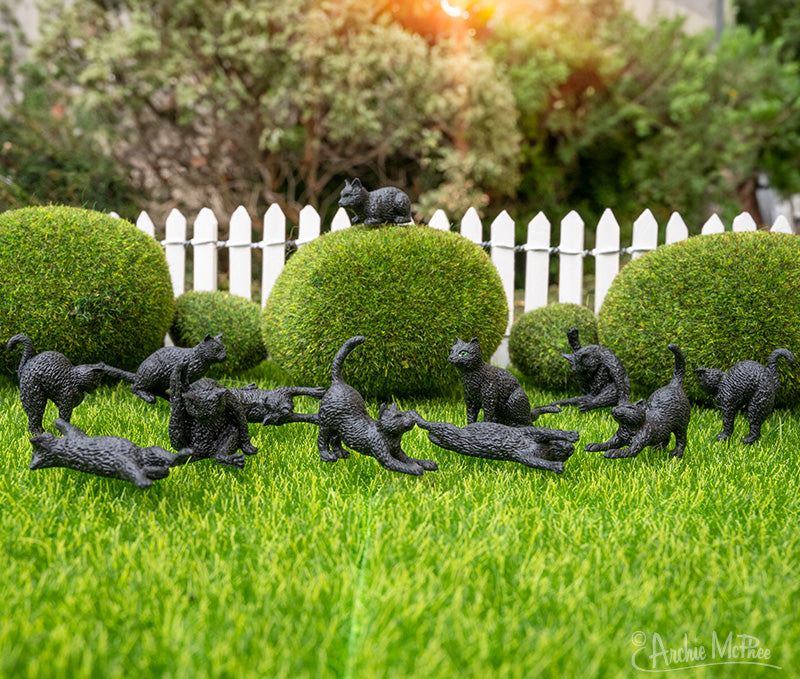 Miniature black cat figurines arranged playfully on grass near topiary bushes and white picket fence, showcasing various cat poses and sizes for the Itty Bitty Black Cats product