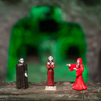 Three miniature vampire figurines in different colored outfits standing in front of a blurred green archway, showcasing the Itty Bitty Vampires product set against a mysterious backdrop