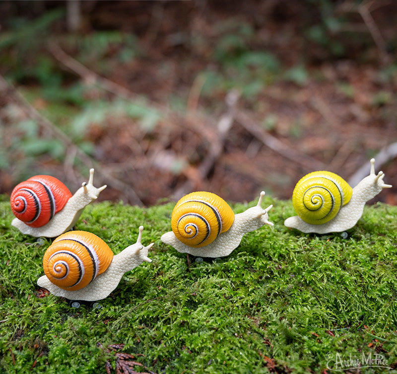 Colorful racing snail toys lined up on moss, featuring vibrant orange, yellow, and red shells with spiral patterns, ready for a playful race in a natural outdoor setting