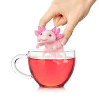 Axolotl-shaped novelty tea infuser being inserted into a clear glass of pink tea by a human hand