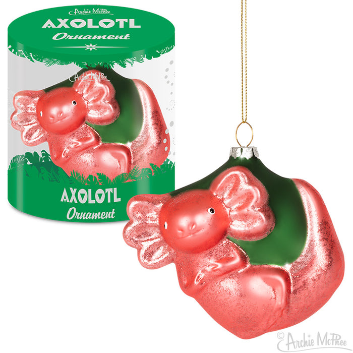 A bright pink axolotl-shaped Christmas ornament with frilly gills, displayed both inside a green cylindrical package and hanging freely. The ornament features a smiling face and curled body, designed to bring unique holiday cheer.