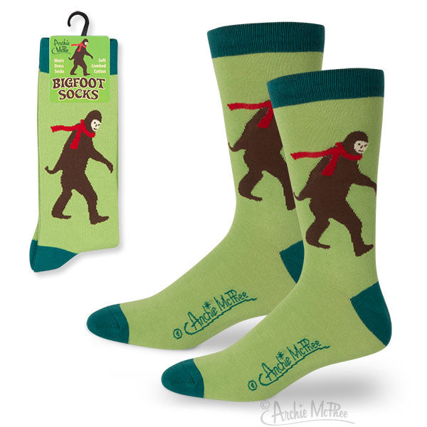 Green men's dress socks featuring brown Bigfoot silhouettes with red scarves, displayed alongside product packaging with matching design. Socks have dark green cuffs and heels, with visible branding on the foot.