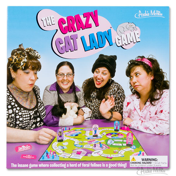 Four women dressed in cat-themed clothing play "The Crazy Cat Lady Game" board game, featuring colorful game pieces and a cat-collecting theme. The box art displays the game title in vibrant letters against a blue background.