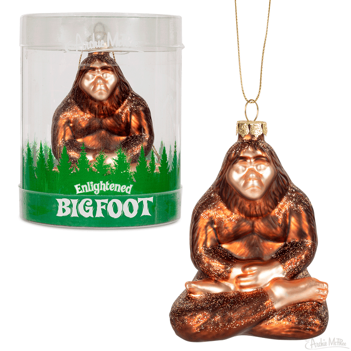 Glass Enlightened Bigfoot Christmas ornament in meditative pose, bronze-colored, seated cross-legged with hands in lap. Displayed in clear cylindrical packaging with green forest silhouette base. Gold string for hanging visible.