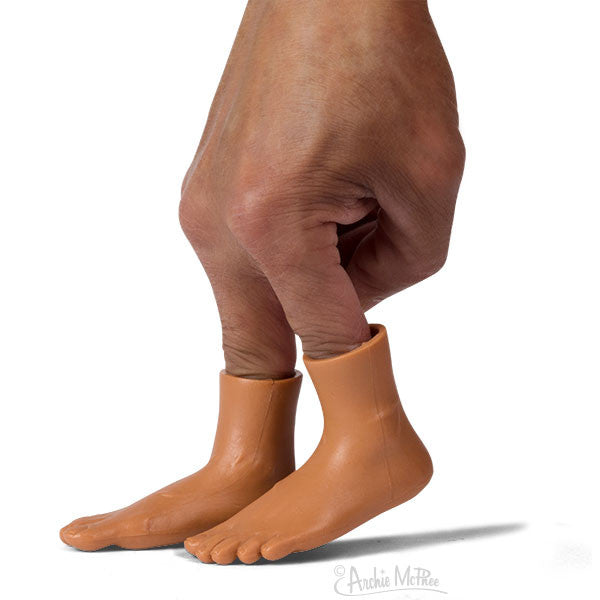 Hand with two fingers wearing miniature vinyl feet in dark skin tone, creating illusion of tiny legs walking on a surface, showcasing Finger Feet product for playful finger transformation