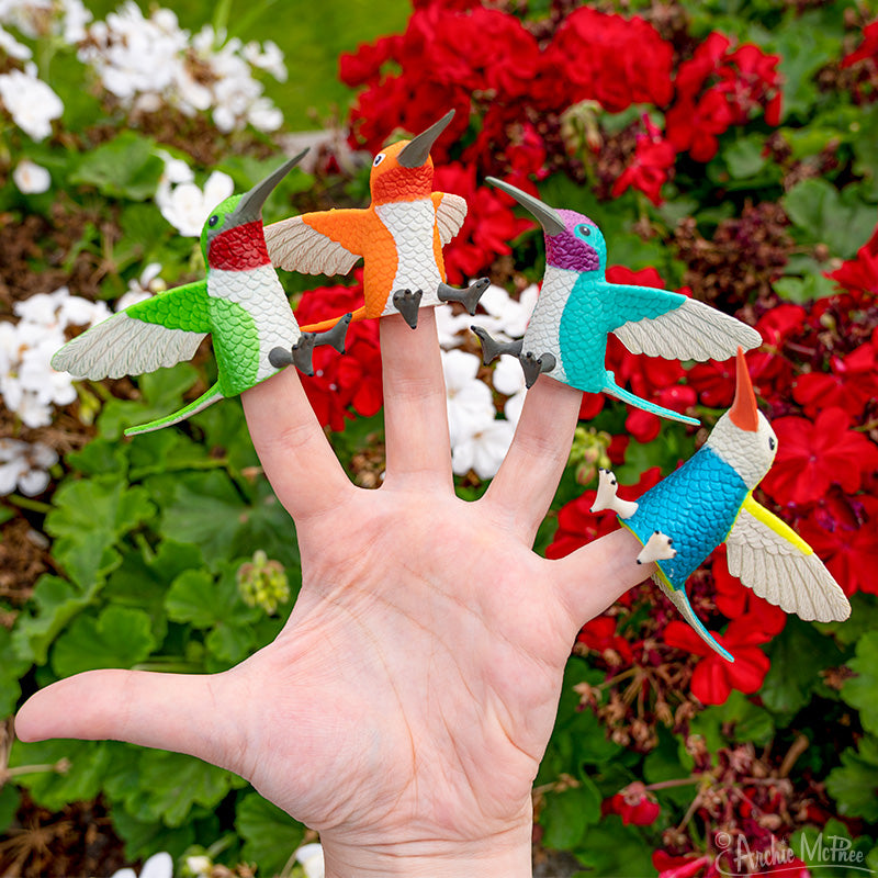 four colorful hummingbird finger puppets on a hand against flowers