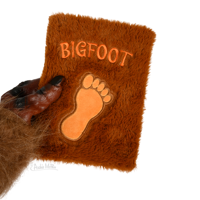 Fuzzy brown notebook with "BIGFOOT" text and footprint design, held by a furry hand, simulating Bigfoot's appearance for a unique and tactile writing experience.