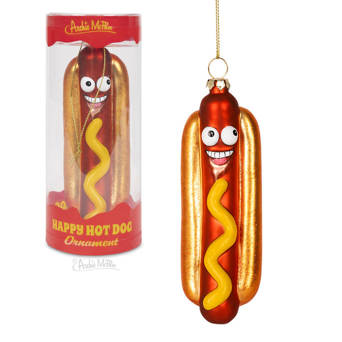 Festive glass hot dog ornament with cartoonish eyes and smile, featuring a golden bun, red sausage, and yellow mustard swirl. Packaged in a cylindrical container labeled "Happy Hot Dog Ornament" by Archie McPhee.