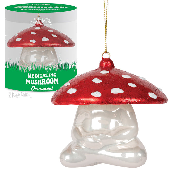 Glass mushroom ornament with red spotted cap and white stem in meditation pose, displayed in green-topped packaging and shown hanging with golden string