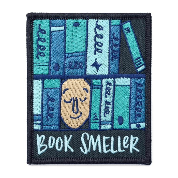 Embroidered patch featuring stylized book spines in shades of blue and teal, with a smiling face in the center. Text at bottom reads "BOOK SMELLER" in white stitching. Square patch with black border.