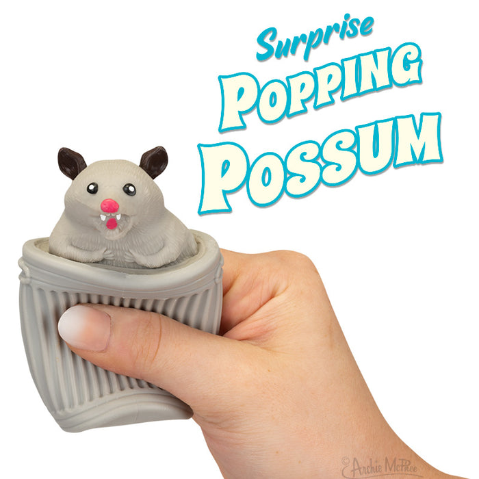 Surprise Popping Possum toy with cute gray possum emerging from a small trash can-shaped container held in a hand, featuring product title in playful blue text