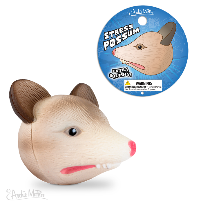 Stress Possum toy with packaging, featuring a beige foam possum head with pink nose and mouth, alongside circular product label showing possum face and 'Stress Possum' text on blue background