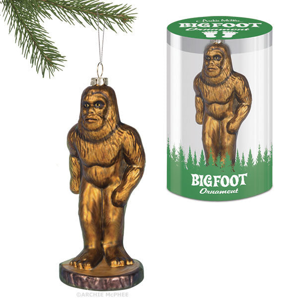 Golden Bigfoot Christmas ornament hanging from pine branch next to product packaging featuring forest silhouette and Bigfoot illustration