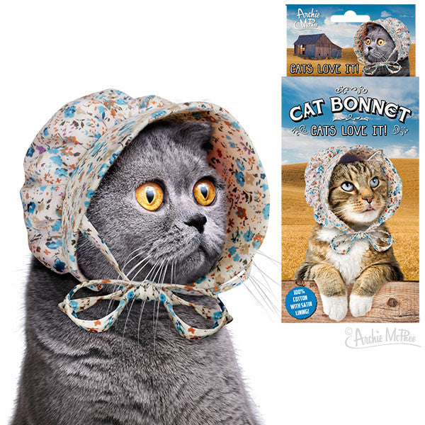 Gray cat wearing floral cotton bonnet with satin lining. Product packaging shows another cat in bonnet against desert background. Text reads "Cat Bonnet" and "Cats Love It!" Unique pet accessory for playful or old-fashioned cat owners.