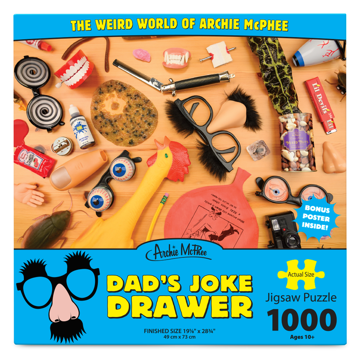 Colorful 1000-piece jigsaw puzzle box featuring "Dad's Joke Drawer" by Archie McPhee, showcasing various novelty items and practical joke supplies on a wooden background, including chattering teeth, whoopee cushion, fake glasses, and rubber chicken.