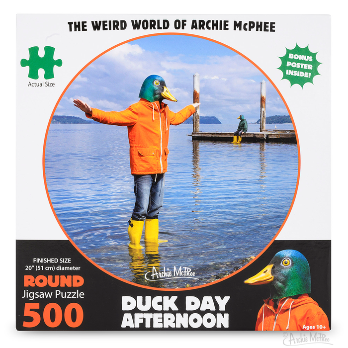 Round jigsaw puzzle box featuring a person in an orange jacket and yellow boots standing in water with arms outstretched. Background shows a dock and calm lake. Puzzle titled "Duck Day Afternoon" with 500 pieces and 20" diameter.