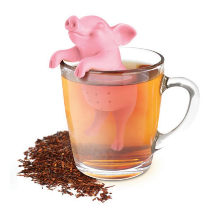 Pink pig-shaped tea infuser sitting on edge of glass mug filled with brewed tea, surrounded by loose tea leaves on white background