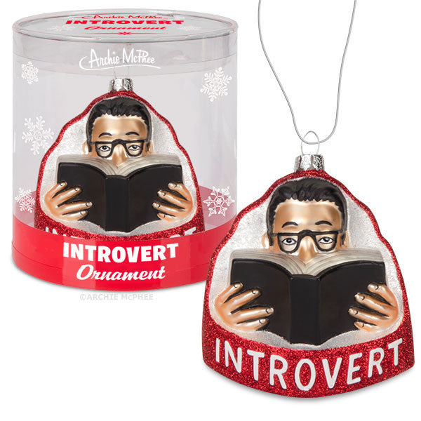 Introvert Ornament featuring a figure reading a book, packaged in a clear cylindrical container with snowflake designs and red accents. The ornament depicts a person with glasses immersed in reading, embodying the introvert theme.