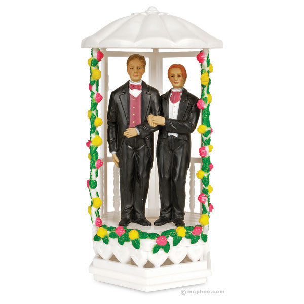 Two groom figurines in tuxedos standing arm-in-arm inside a white gazebo decorated with colorful floral garlands. Cake topper for same-sex wedding celebrations.