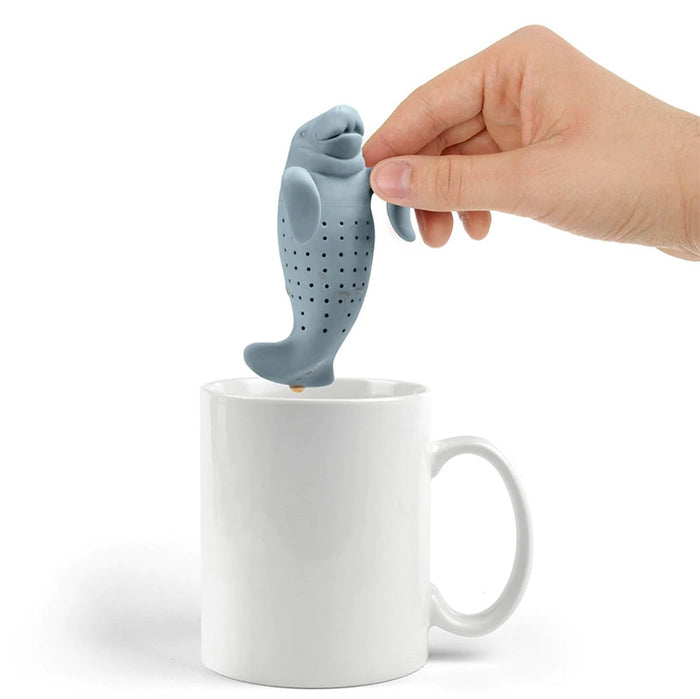 Hand holding a Manatea Tea Infuser, a gray silicone manatee-shaped tea infuser with small holes, being lifted from a white ceramic mug. The unique, playful design showcases the product's functionality for steeping loose leaf tea.