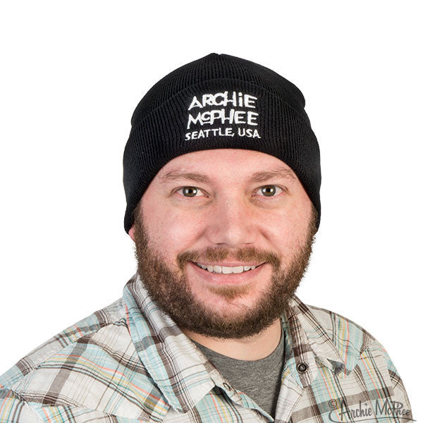 Black knit beanie with "ARCHIE MCPHEE SEATTLE, USA" embroidered in white, worn by a smiling person in a plaid shirt against a white background.