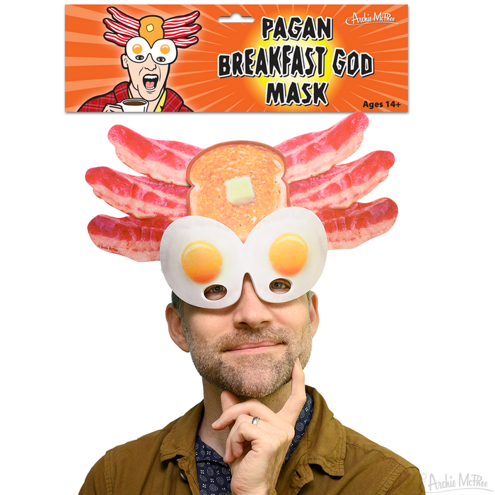 Man wearing quirky breakfast-themed mask featuring toast, bacon strips, and fried eggs, with product packaging showing "Pagan Breakfast God Mask" text and cartoon character