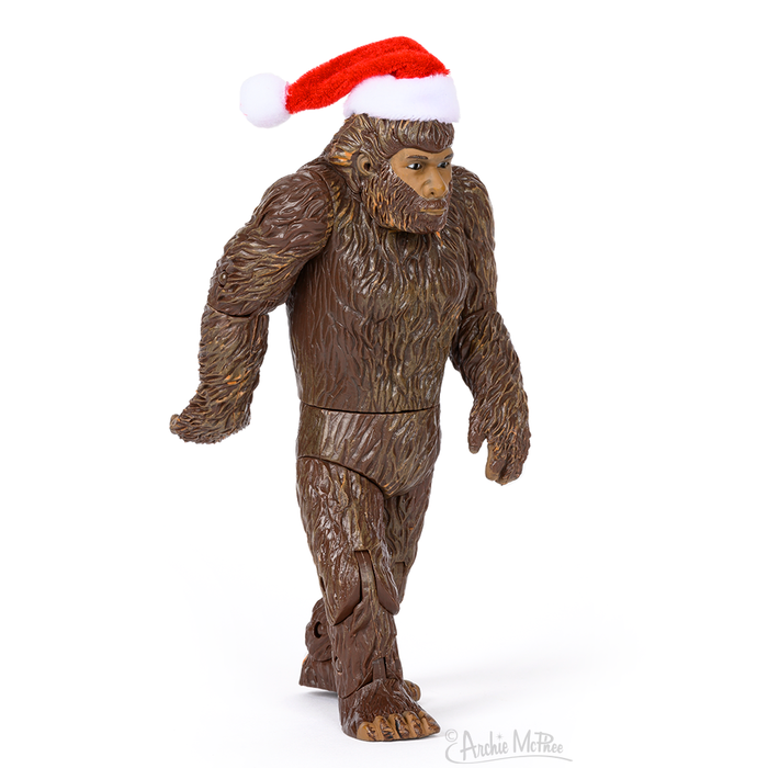 Santa Bigfoot action figure with brown fur, wearing a red and white Santa hat, standing upright in mid-stride pose, resembling the famous Patterson film footage. Articulated vinyl toy perfect for Christmas decorations or Sasquatch enthusiasts.