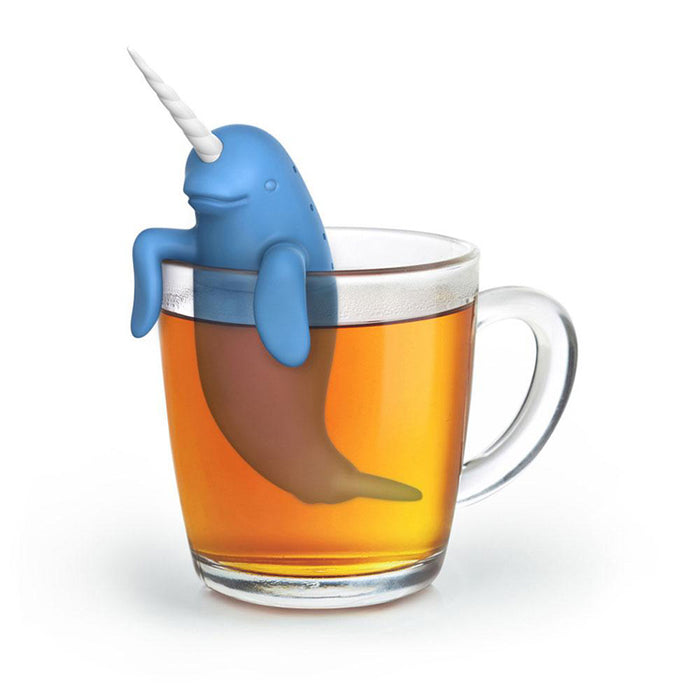 Cute blue narwhal-shaped tea infuser perched on the rim of a glass mug filled with golden tea, its long tusk extending upwards and tail submerged in the beverage