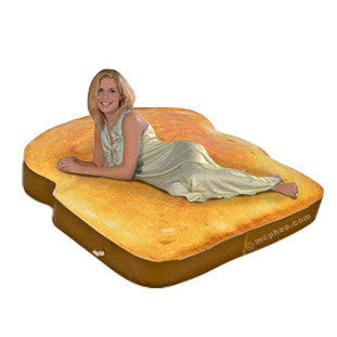 Inflatable mattress shaped like a giant slice of toast, golden-brown in color with a raised crust edge. A woman in a light-colored dress is lying on the mattress, demonstrating its size and comfort. The novelty bed appears soft and inviting.